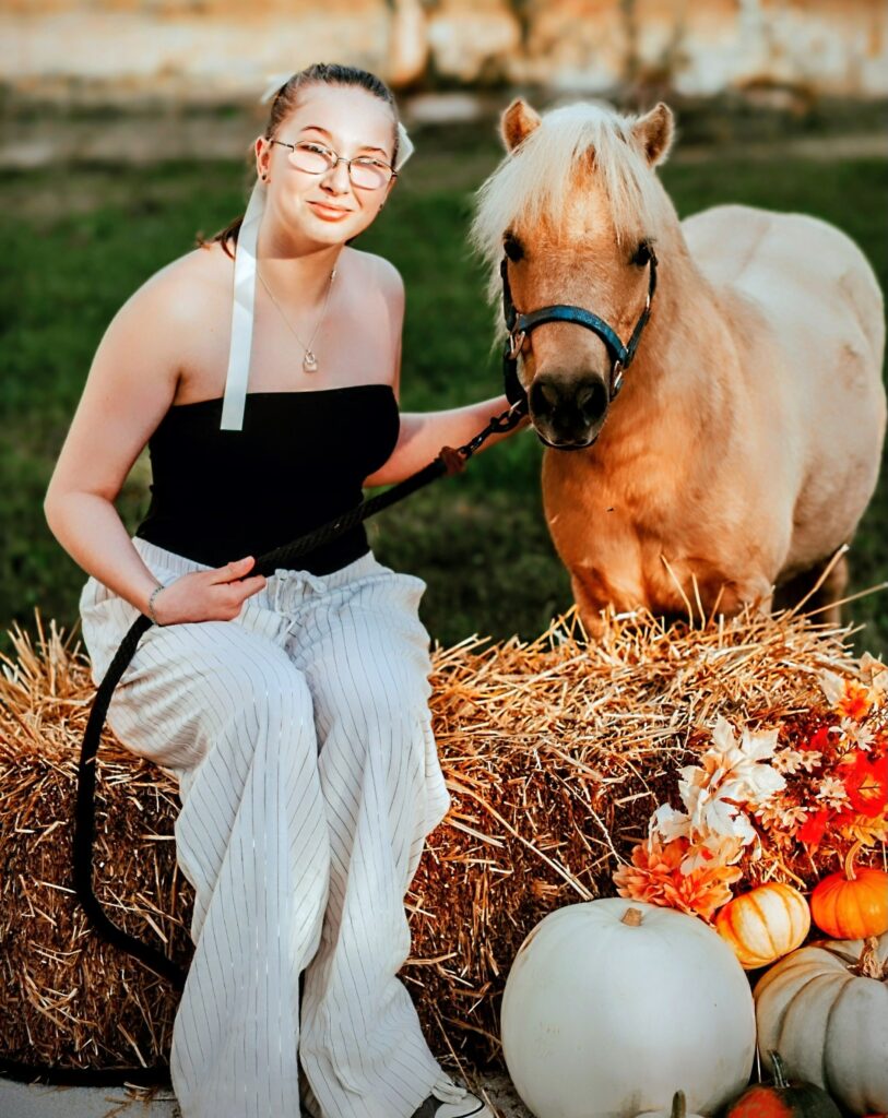 High school student sits on a bale of straw while holding the lead rope of a palomino colored miniature horse.