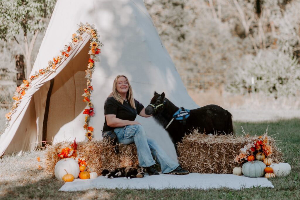 Founder of Equine Eyes sits on a bale of straw next to Mijo, a blue roan miniature guide horse prospect.